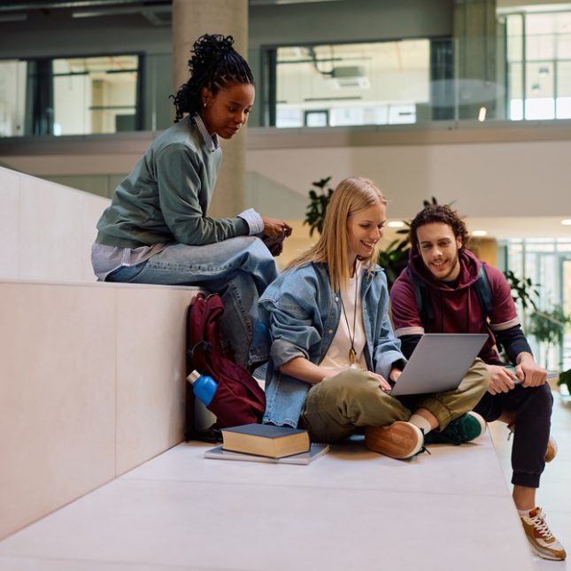 Grupa studentów uczy się wspólnie, siedząc na trawie z laptopami i notatkami